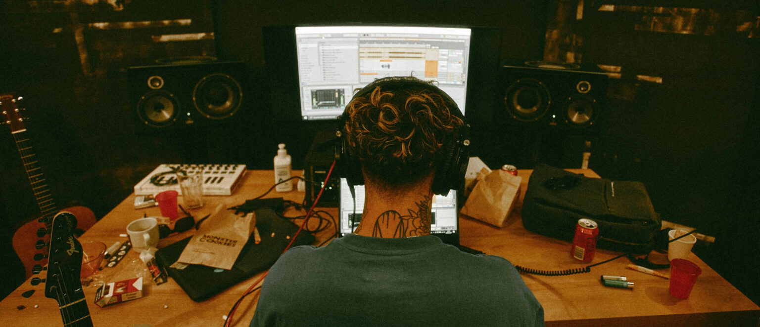 A creator sitting in their studio facing their computer and monitor speakers. Two guitars can be seen propped up next to their desk.